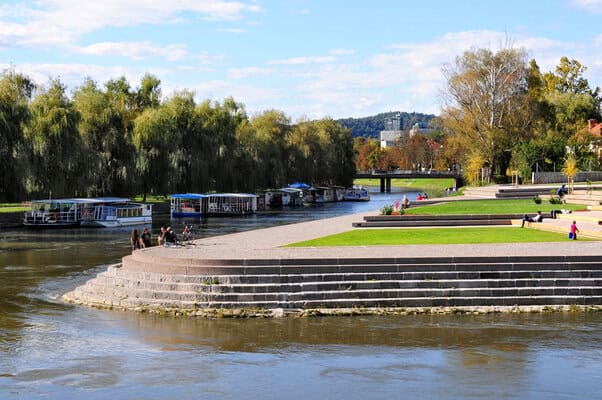 Scenic view of Ljubljana River with boats, lush trees, and walking paths in Ljubljana, Slovenia.