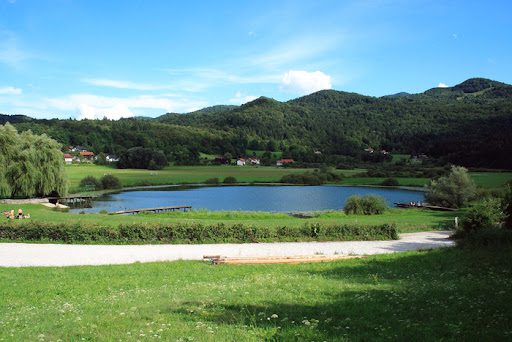 Podpec Lake, a natural lake near Ljubljana, Slovenia, surrounded by lush green meadows, forested hills, and wooden jetties on a clear summer day.
