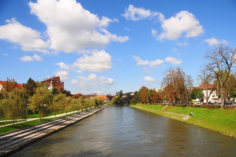 View of the Ljubljanica River flowing between tree-lined banks, showing the stone-stepped Trnovski Pristan aka beach on the left and grassy banks on the right.