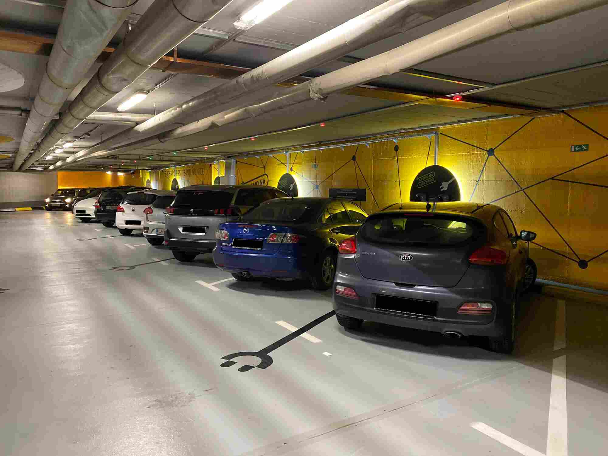 Designated electric vehicle charging bays within the modern Šubičeva underground parking garage (PH Šubičeva) in Ljubljana, Slovenia, marked by bright yellow walls and EV floor symbols.
